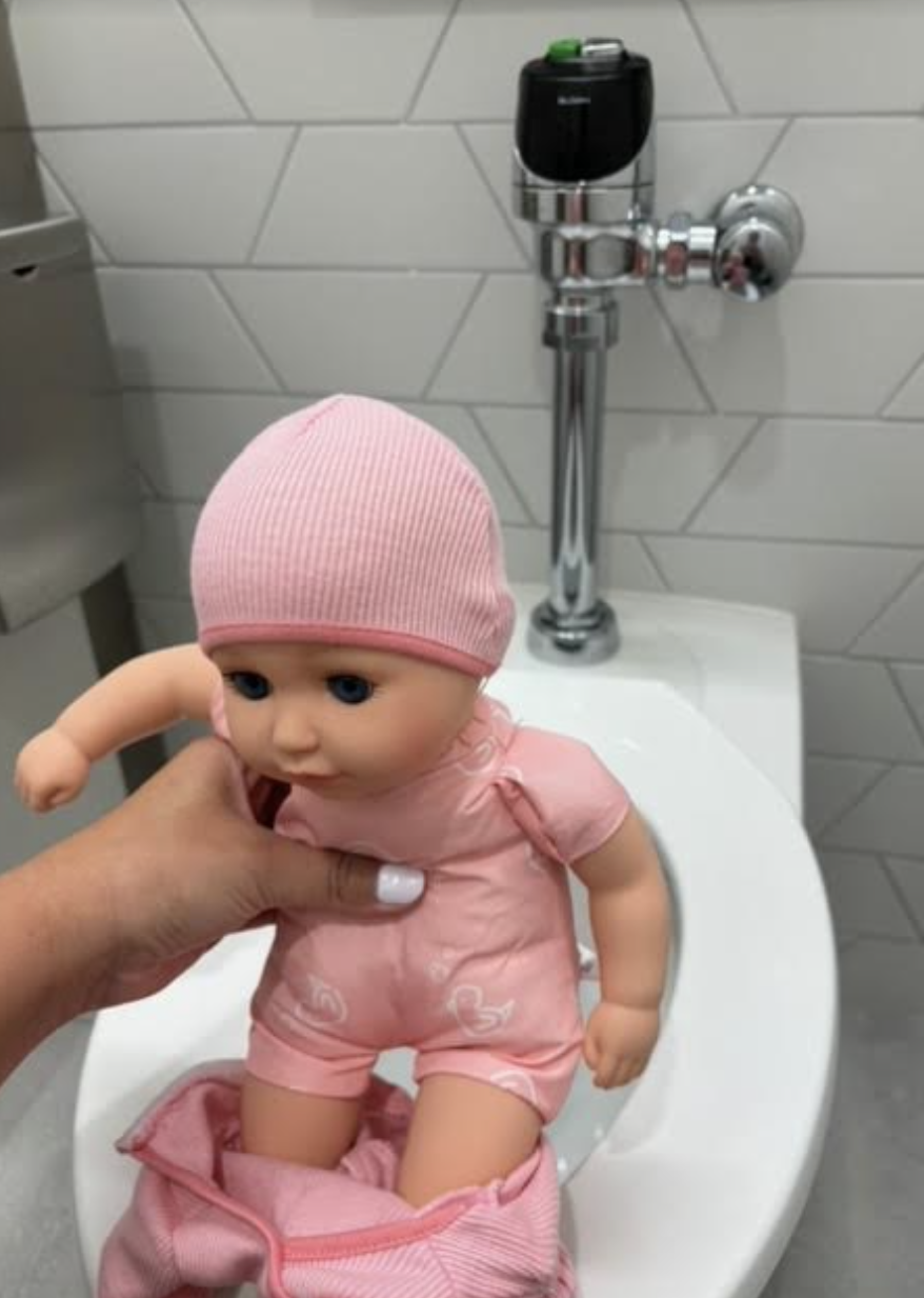 A hand is holding a baby doll dressed in a matching hat and outfit over a toilet