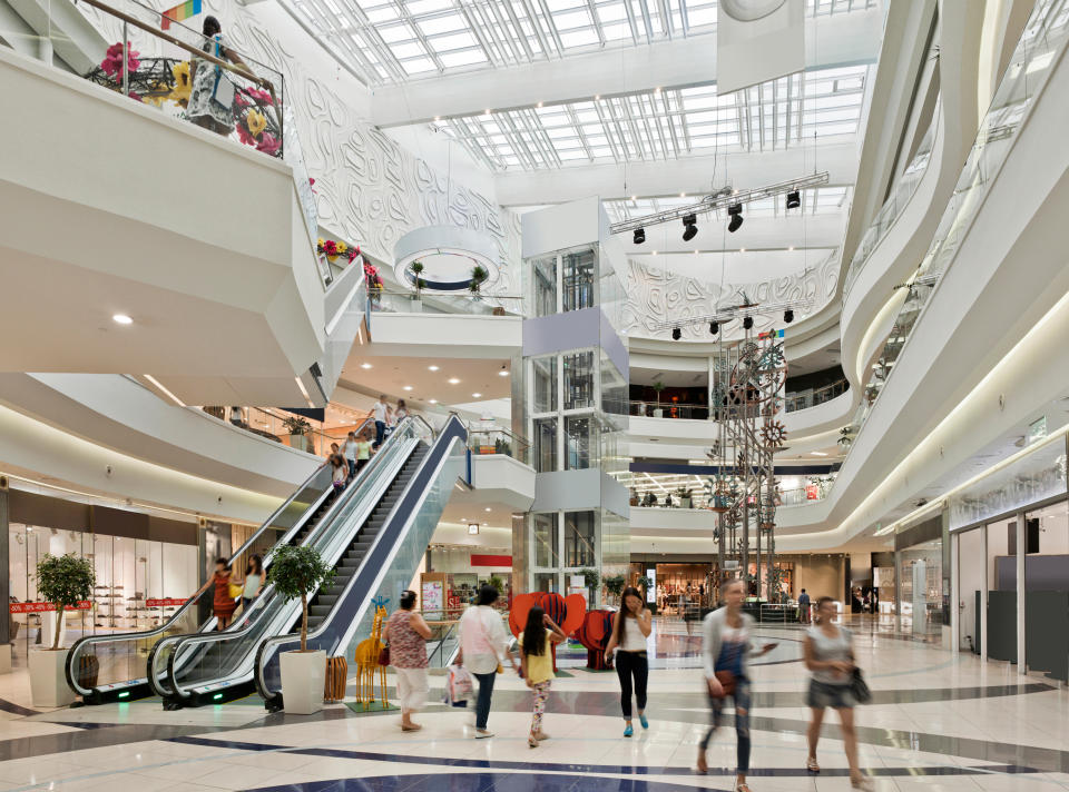 People walking on the ground floor of a spacious, modern shopping mall with shops, escalators, and a large sculpture in the center