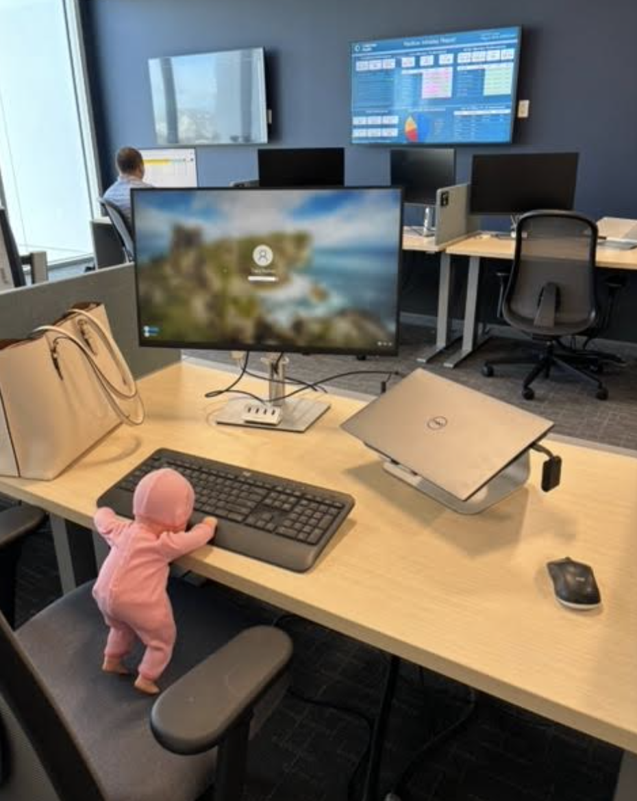 A baby doll in a pink outfit is propped up against a keyboard on a desk in an office setting. Multiple desks and monitors are visible in the background