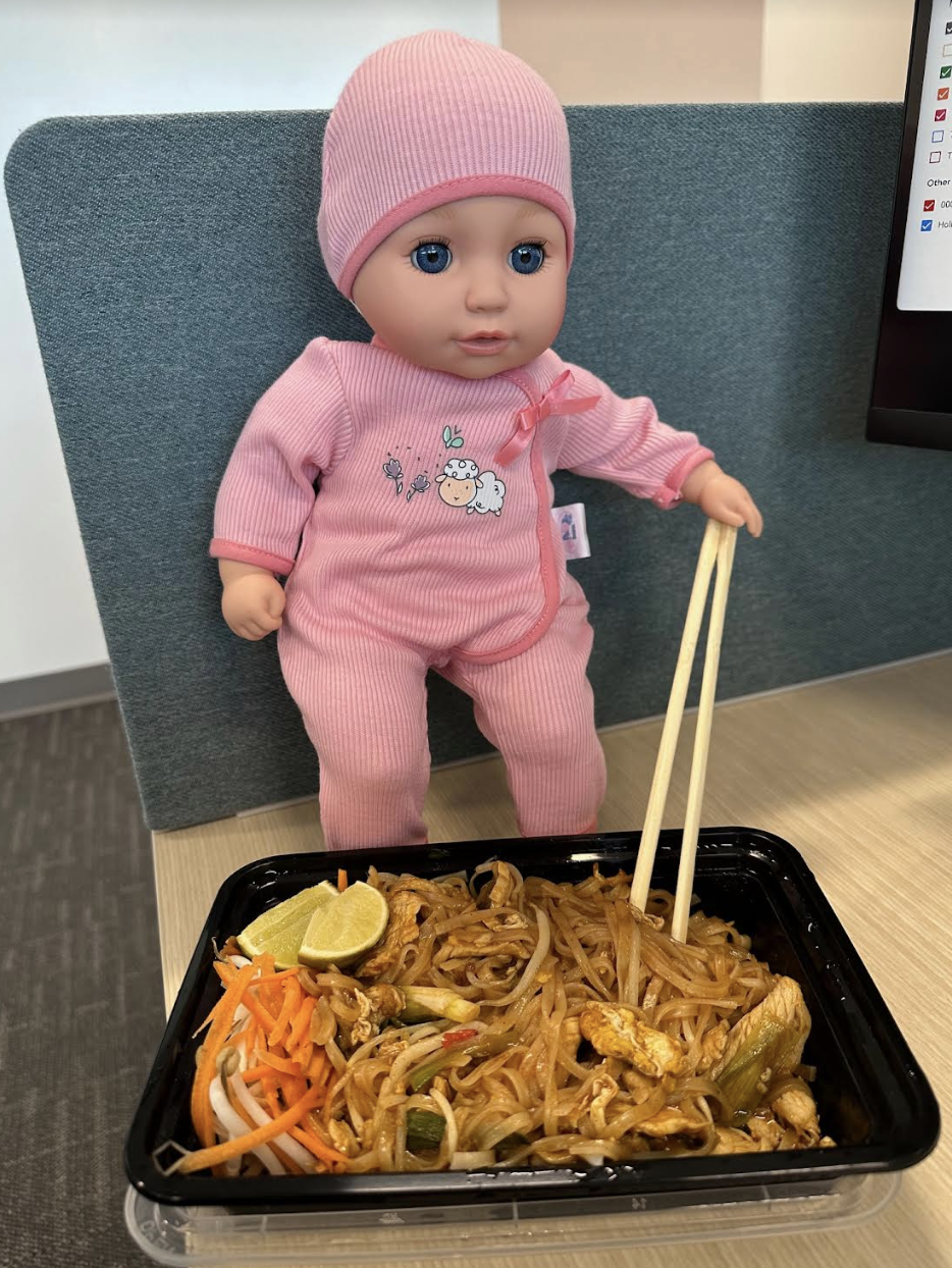 A baby doll wearing a onesie and hat is leaning on a table with chopsticks, facing a container of noodles with vegetables, lime, and chicken