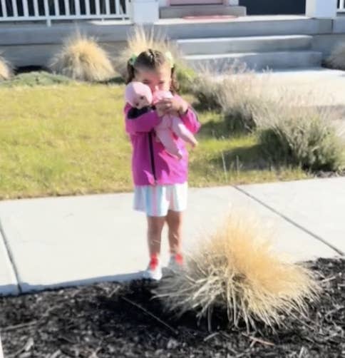 Young girl holding a stuffed animal and standing on a sidewalk with grass and plants around. She has pigtails and wears a pink jacket and a skirt