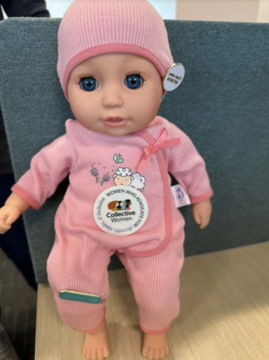 A doll with blue eyes wearing a pink outfit with a sticker that reads "Women Who Advocate" and "Collective Women."