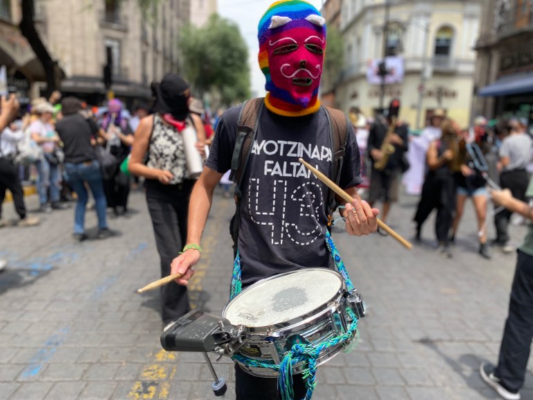 A man in a knitted full-face mask beats a drum as he marches. His homemade T-shirt reads: Ayotzinapa 43.