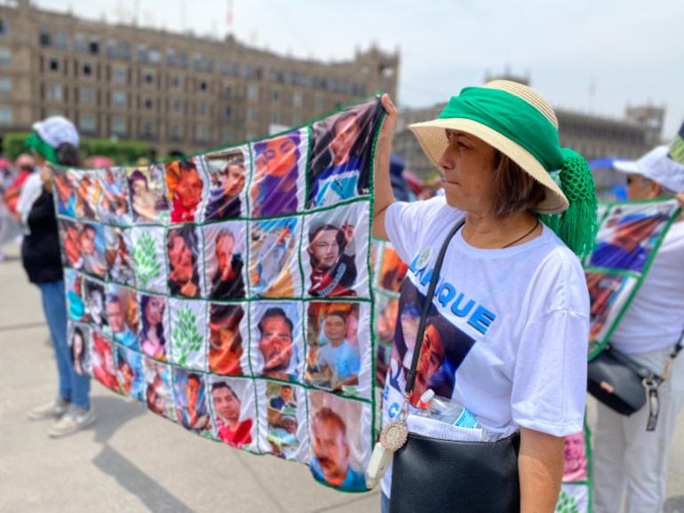 A woman in a straw hat holds up one side of a large banner that shows the faces of missing people in Mexico. Her T-shirt is likewise printed with the face of a man, Enrique, who has gone missing.