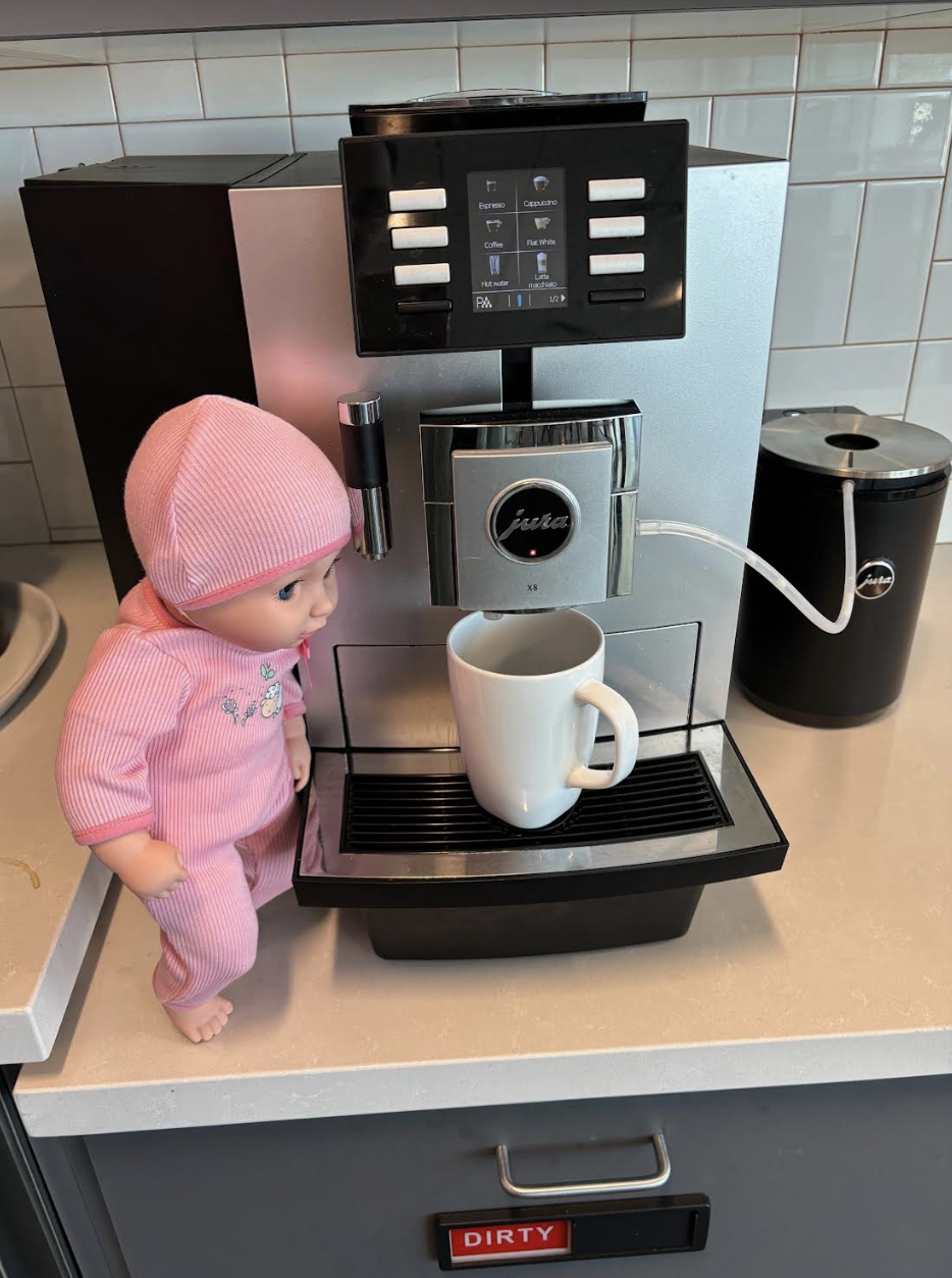 A baby doll dressed in pink pajamas stands next to a Jura coffee machine with a white mug placed under the coffee spout