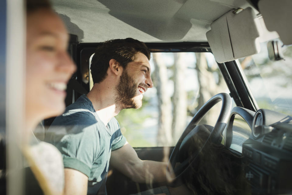 Two smiling people sit in a car. The person on the left is blurred, while the person on the right is clearly visible, driving the car and laughing