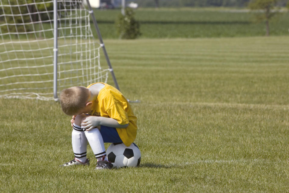 A boy in a soccer uniform sits on a soccer ball on the field, looking down with his head in his hands. The goal net is in the background