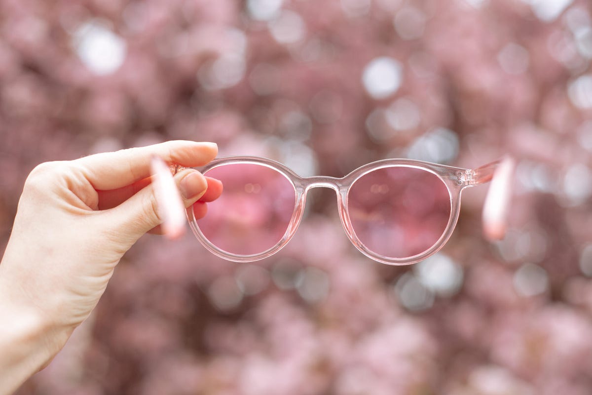 Hand holding pink sunglasses against a blurry pink background.