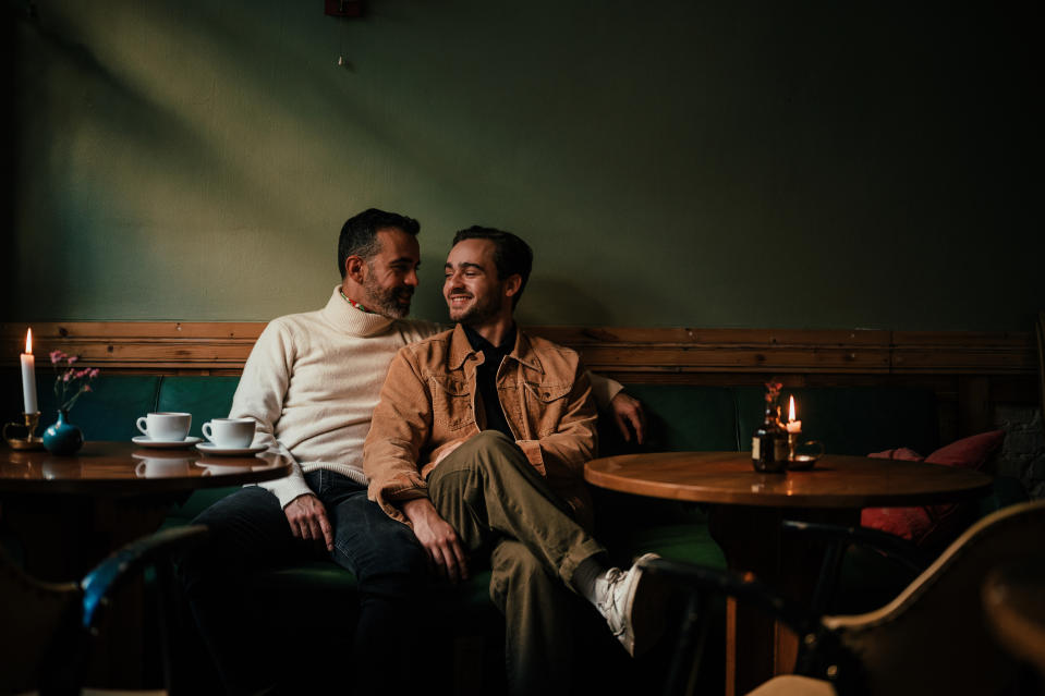 Two men sitting closely on a cozy bench in a dimly lit café, smiling at each other with cups of coffee on the table