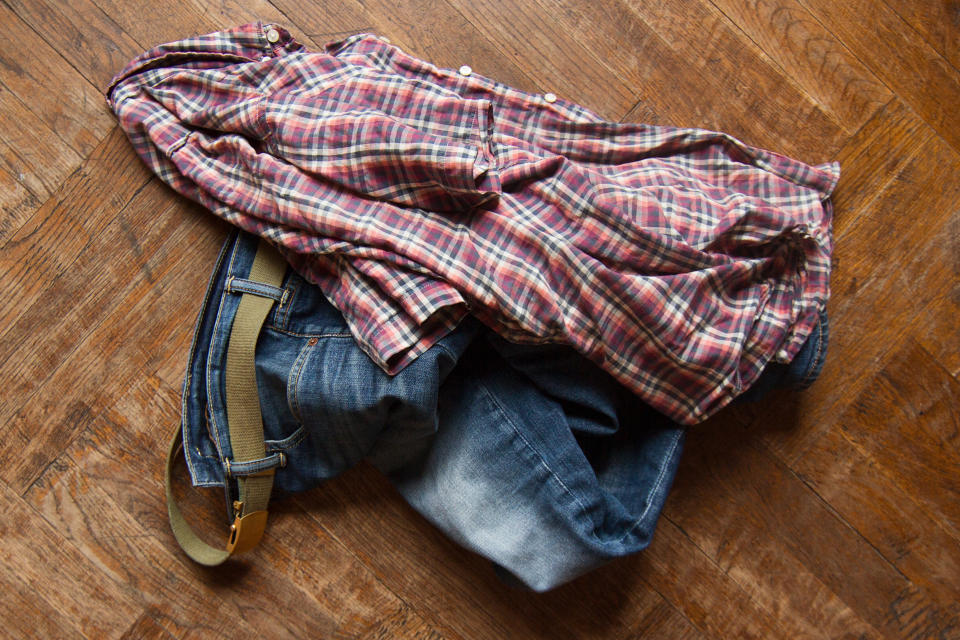 A crumpled plaid shirt and blue jeans with a belt lie on a wooden floor