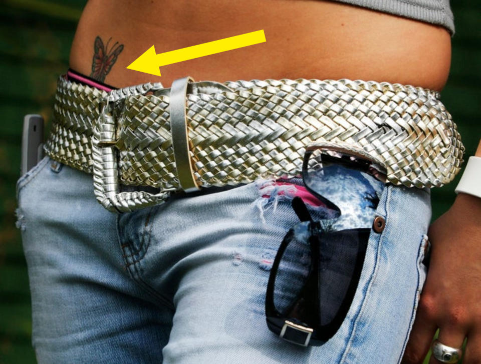 A close-up of a person's waist wearing blue jeans, a thick woven belt, and sunglasses hanging from the jeans pocket. A butterfly tattoo is visible above the jeans