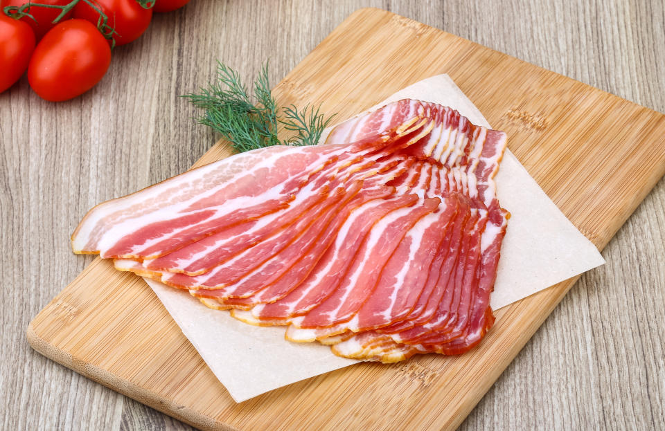 Slices of bacon neatly arranged on a wooden cutting board, with a few sprigs of dill and tomatoes in the background. The image is categorized under Sex & Love