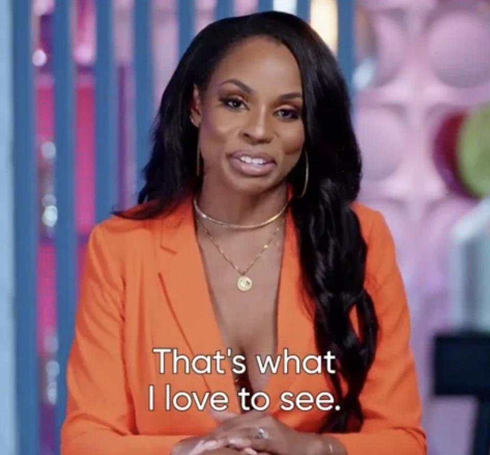 A woman with long dark hair, wearing an orange blazer over a low-cut top and a necklace, is shown speaking. Text on image reads, "That's what I love to see."