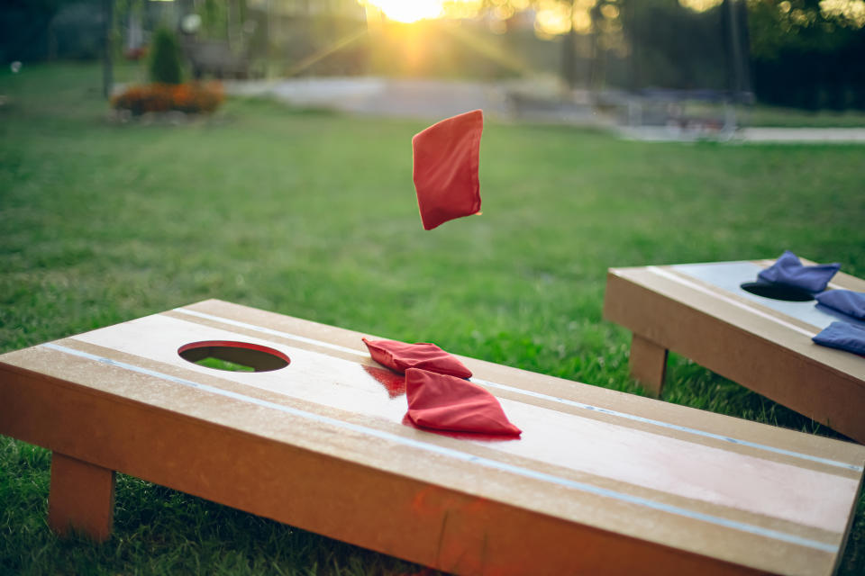 Two cornhole boards on grass, with red and blue bean bags mid-game as the sun sets in the background. Focus on a red bean bag in motion towards the hole