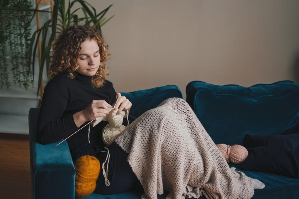 Person sitting on a couch, knitting with yarn, covered with a blanket. Houseplant in the background. Peaceful, cozy setting