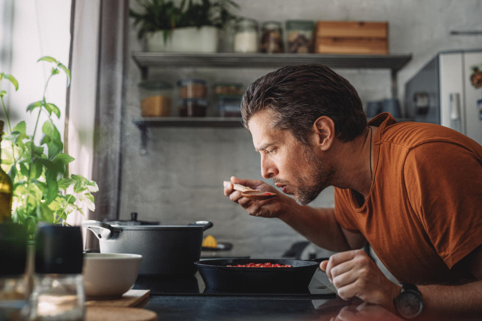 Man in kitchen, tasting dish from spoon, standing by stove with pots and pans