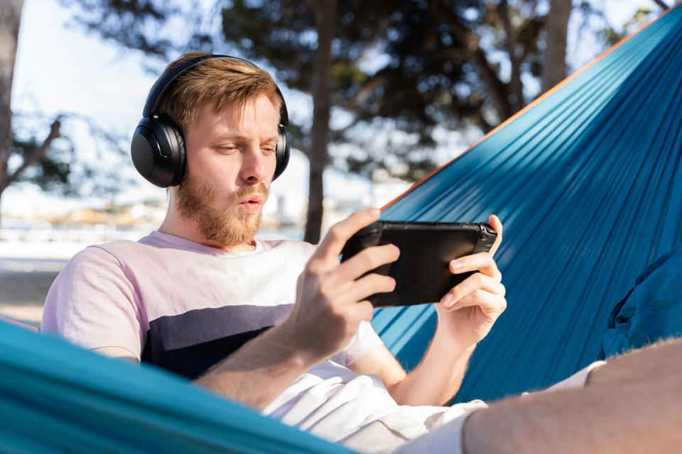 Man in a hammock wearing headphones and reacting to something on his tablet, outdoors