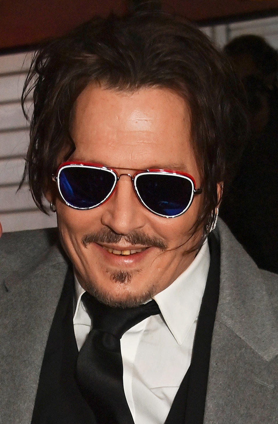 Johnny Depp in a grey blazer, white shirt, black vest, and tie, wearing reflective aviator sunglasses with a red and blue frame, smiling for the camera
