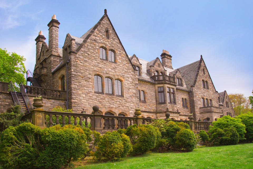 Large stone mansion with multiple gables and chimneys, surrounded by manicured bushes and a lush lawn