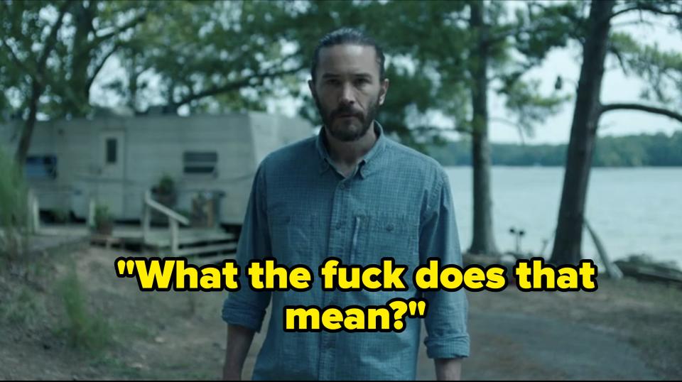 A man with a beard and ponytail, wearing a button-up shirt, stands outside near a lake and a trailer. Trees and water are visible in the background