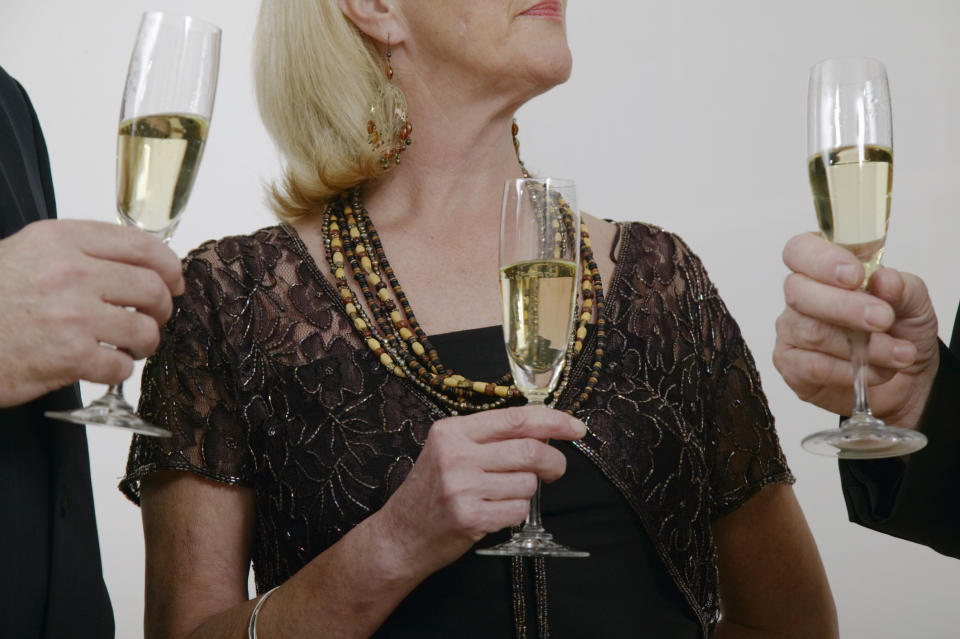 Three people, one elderly woman and two others, hold champagne glasses in a celebratory setting. The elderly woman wears a beaded necklace and lace top