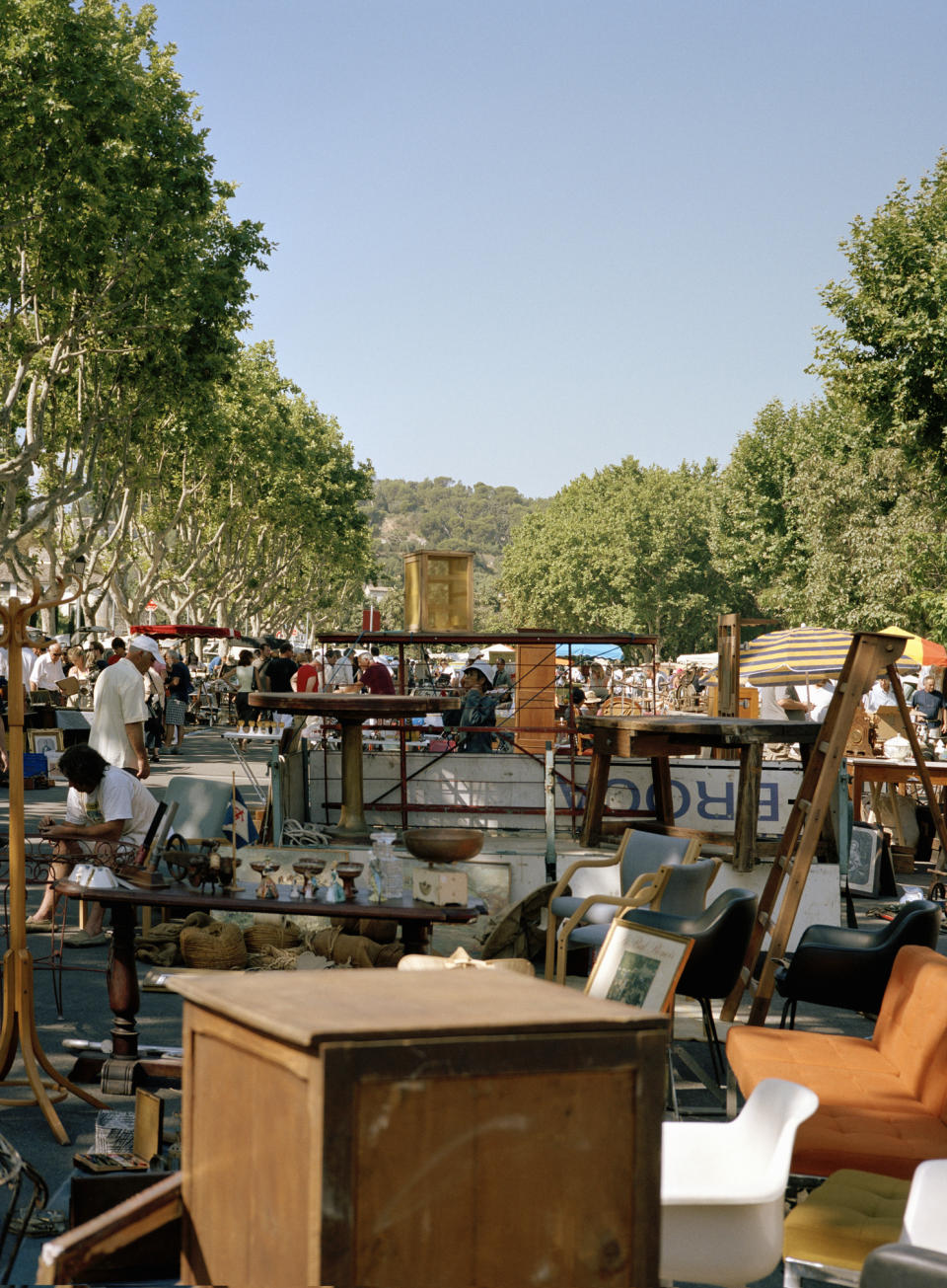 Busy outdoor flea market with various furniture and items on display, numerous people browsing in the background. Trees line the bustling market area