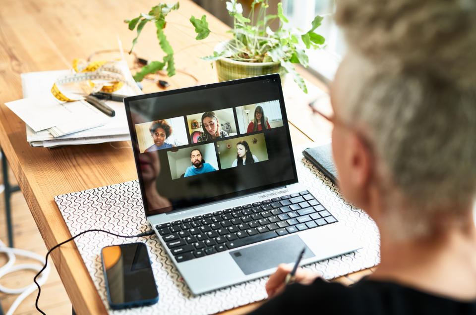 A person participates in a Zoom meeting with five colleagues displayed on the laptop screen. Items like papers, a phone, and a plant are on the desk