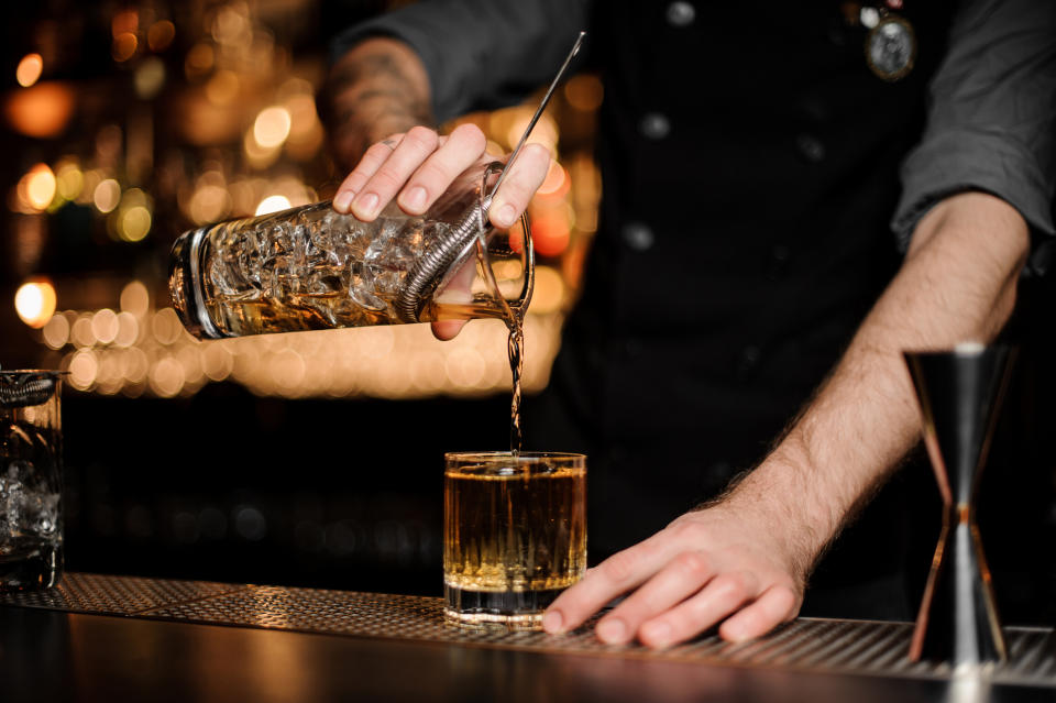 Bartender pouring a drink from a strainer into a glass at a bar. The image illustrates a scene of work in the bartending profession