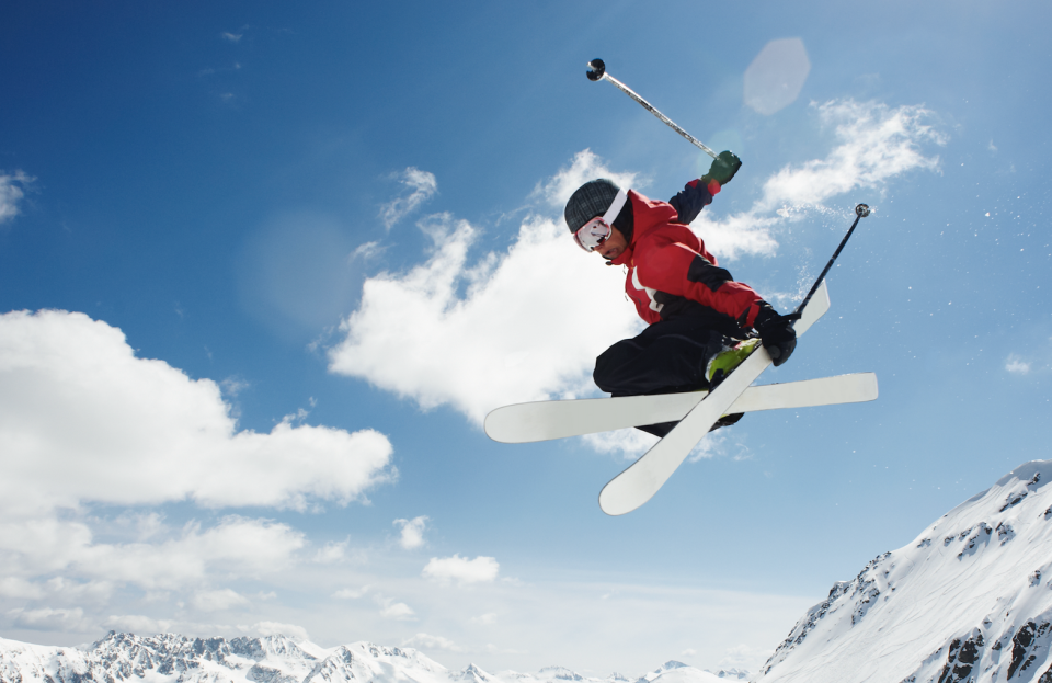 A skier in mid-air performs a stylish jump over a snow-covered mountain range with a clear sky backdrop