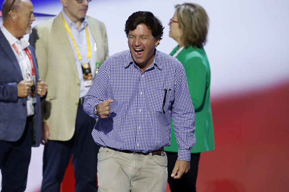 Tucker Carlson laughs while wearing a checkered shirt and tan pants, with three other people in the background