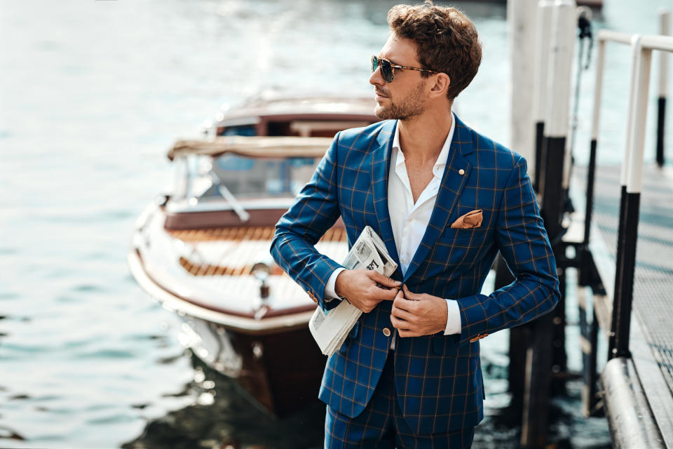 A man in a stylish checked suit and sunglasses stands near a boat on the water, holding a rolled-up newspaper and looking off into the distance