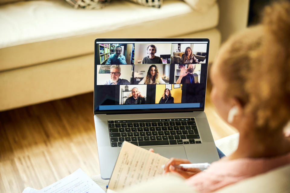 A person attending a virtual business meeting on a laptop from home, holding a notebook and pen, with multiple colleagues visible in the video call