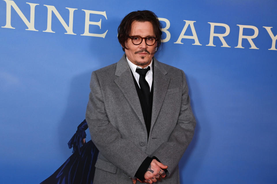 Johnny Depp stands in front of a "Minamata" premiere backdrop, wearing a grey coat over a black suit and tie