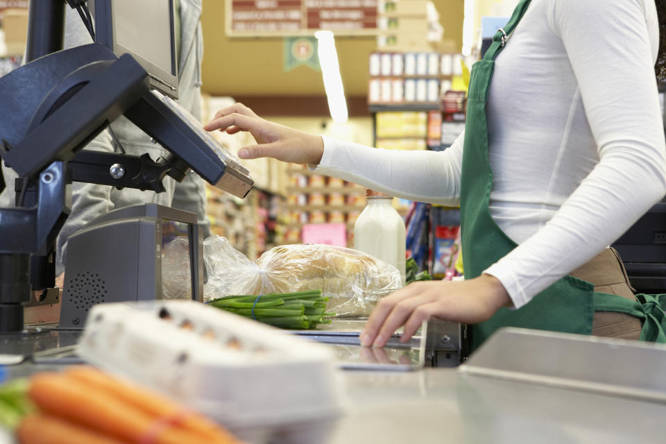 A cashier, wearing a green apron, scans groceries including a loaf of bread, milk, green onions, and carrots at a checkout counter