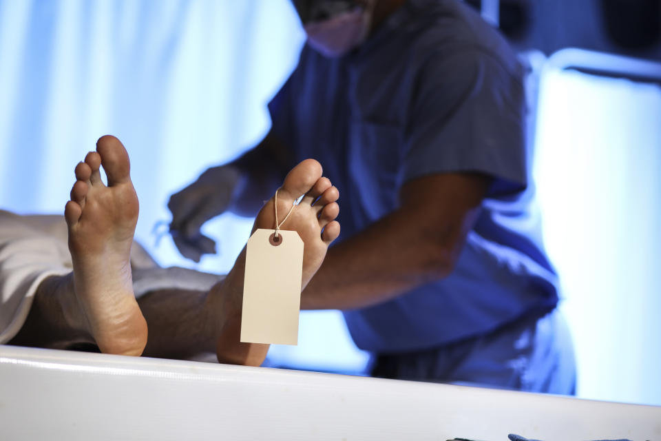 A person's feet on an autopsy table, with a toe tag attached. A medical professional wearing scrubs is seen in the background preparing for examination