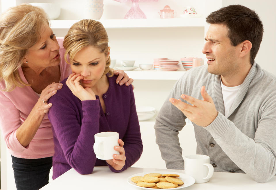 Three people in a kitchen: an older woman comforting a worried younger woman holding a coffee mug, while a man in a sweater gestures intently with cookies on the table