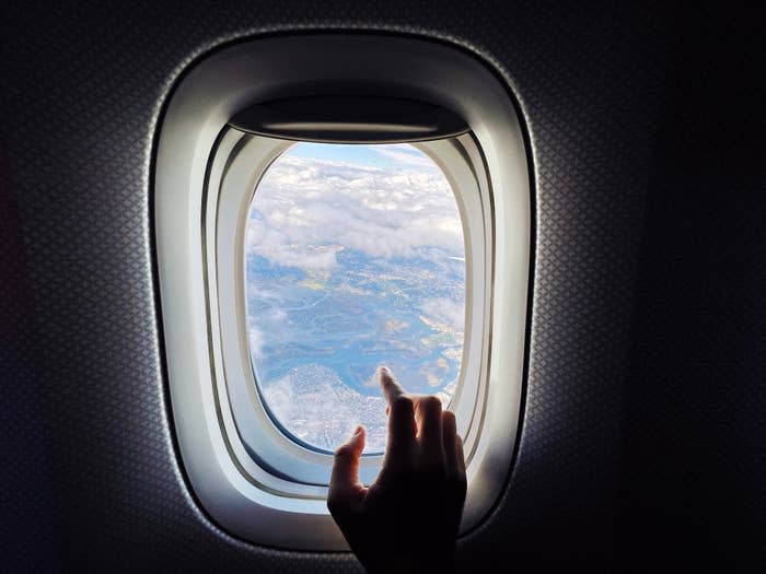 A hand pointing out an airplane window at scenic landscapes of clouds and land below