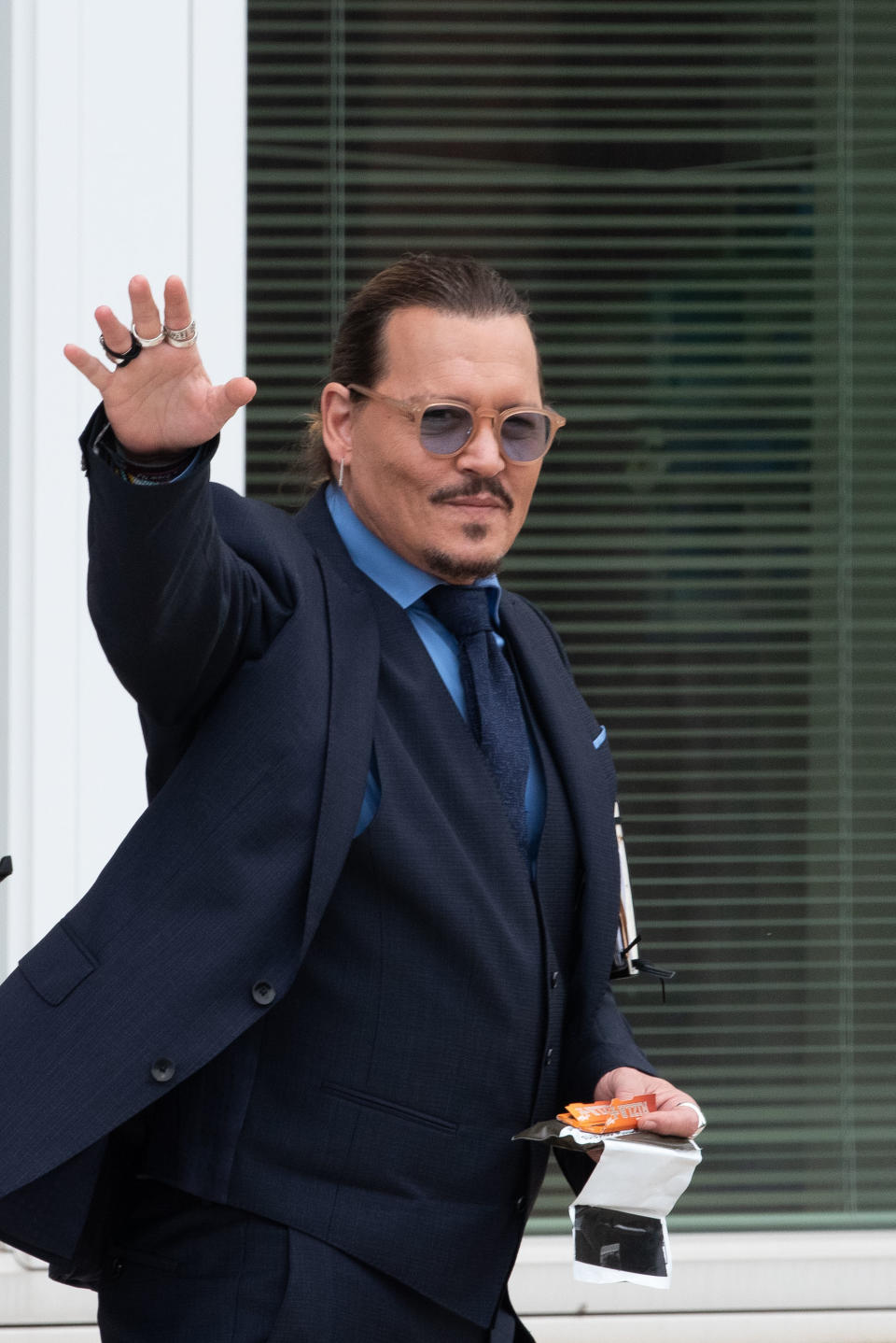 Johnny Depp waves while wearing a dark suit, blue shirt, and sunglasses, holding a small snack and tissues