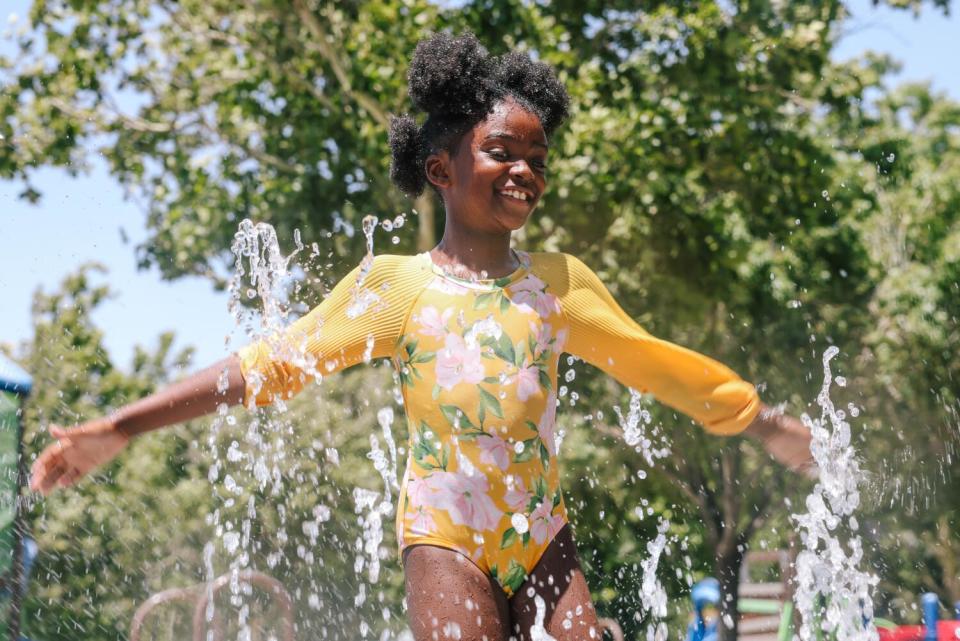 A girl in a yellow swim outfit with a floral pattern extends her arms and smiles while playing in fountain jets at a park