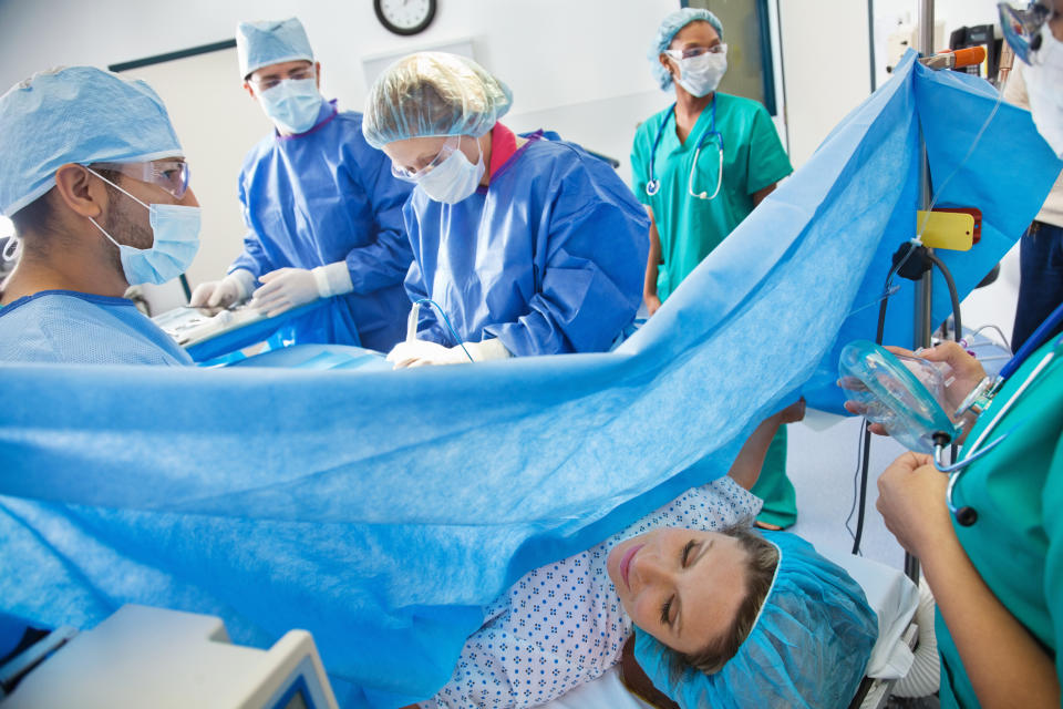 A woman lies on an operating table, partially covered by a surgical drape, while a team of medical professionals performs surgery