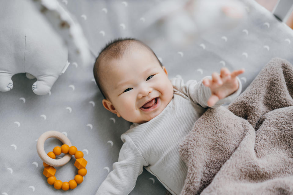 A smiling baby lies on a bed beside a beaded ring toy, reaching out with one hand while partially covered by a blanket. The setting is playful and cozy