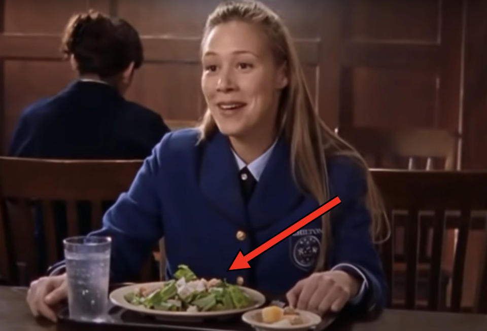 Woman in a school uniform with "CHCETON" badge, smiling while seated at a table with a meal of salad, bread, and a glass of water