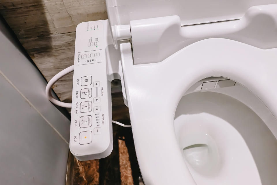 Modern porcelain toilet with a built-in bidet control panel attached to the side. The buttons on the control panel indicate various bidet functions
