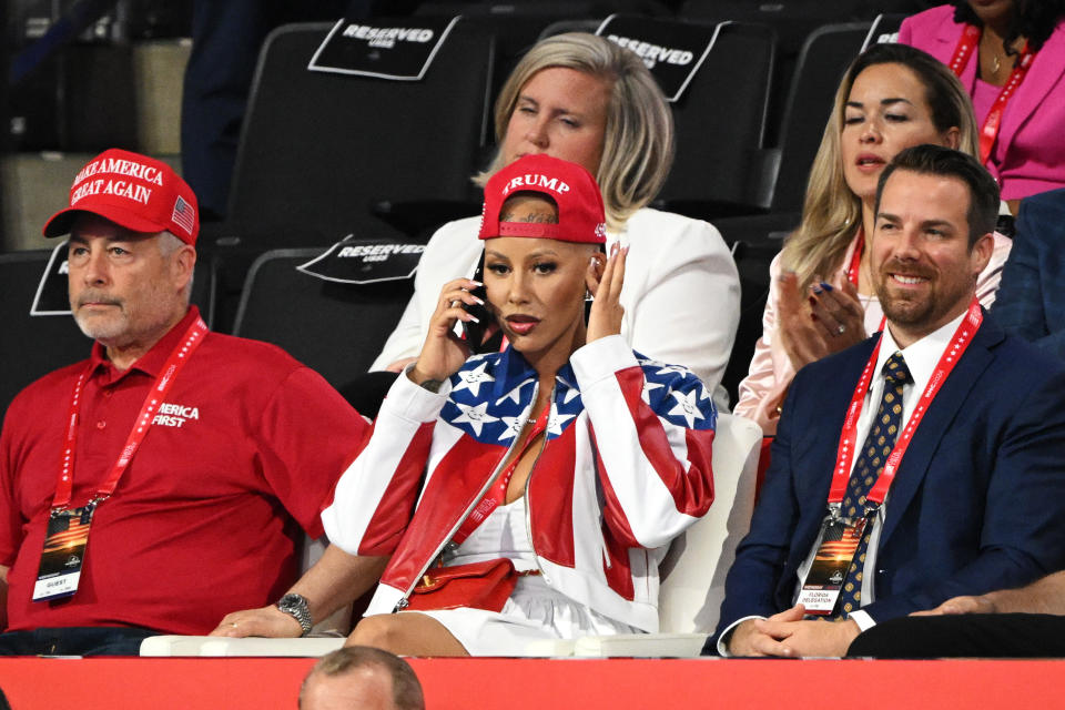 Several people, including Mel B, are seated at an event. Mel B is wearing a patriotic-themed outfit and a red Trump hat while talking on a phone