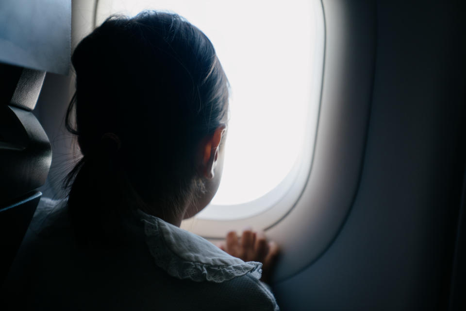 A young child with a ponytail looks out the window of an airplane, hands resting on the window frame