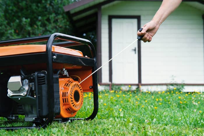 A person's hand pulls the starter cord of an orange and black portable generator in a grassy yard, with a small house in the background