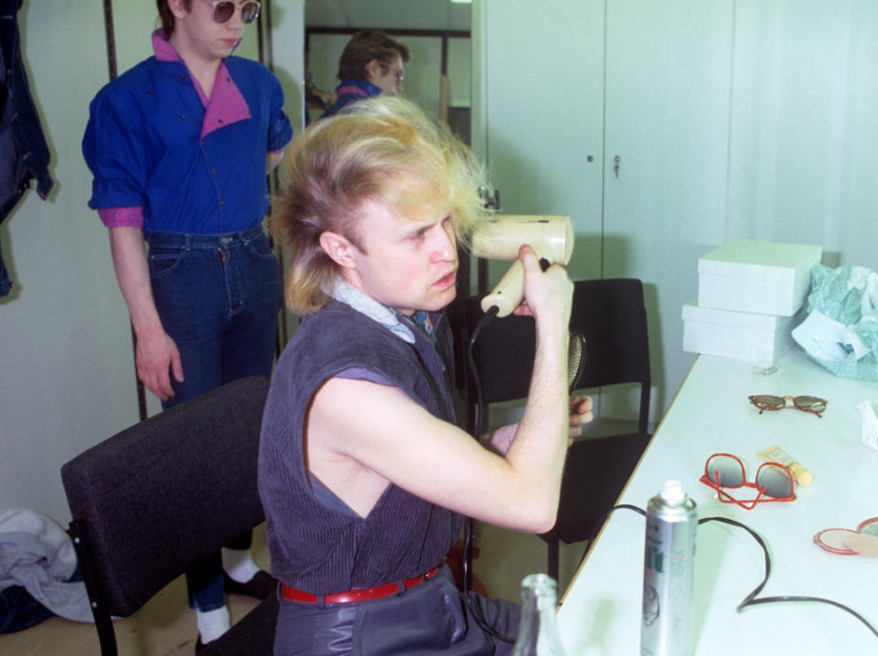 Two people in a backstage dressing room. One, in the foreground, is blow-drying their hair while seated; another stands in the background wearing sunglasses