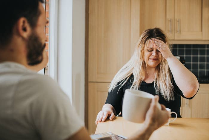 A woman sitting at a kitchen table holds her forehead in distress while a man in the foreground holds a mug. They are having a serious conversation
