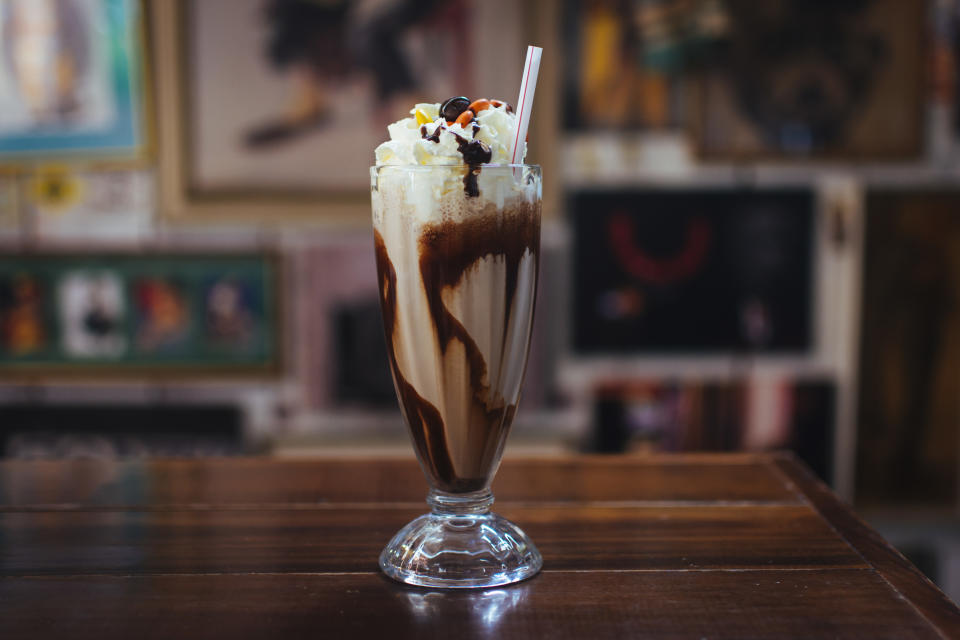 A tall glass of a milkshake topped with whipped cream, chocolate syrup, and a cherry on a wooden table in a decorated room
