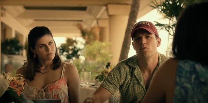 Alexandra Daddario and Jake Lacy are seated at an outdoor table, conversing with a person whose back is to the camera, in a casual setting